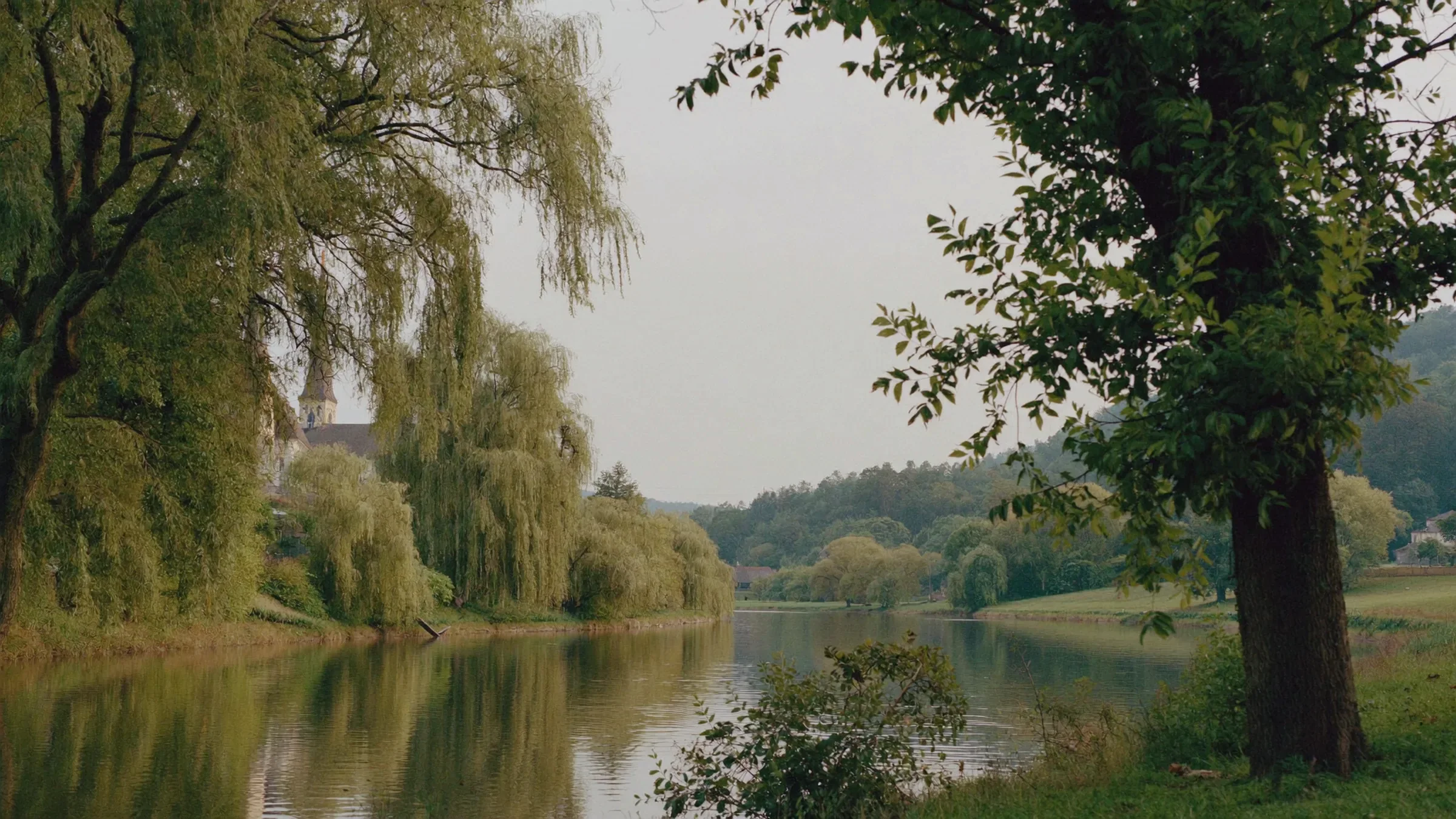 Ruhiger Flusslauf mit Weiden am Ufer, dahinter ein Kirchturm im Morgendunst